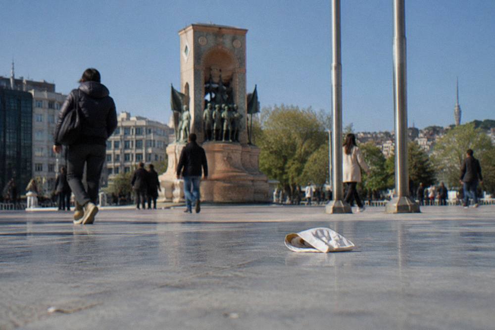 Pedestrian plaza with a stone monument, flagpoles, and crowds walking across an open square