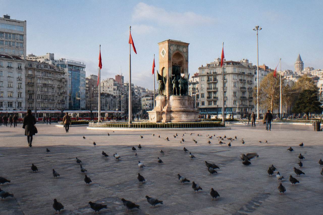 Open urban square with a monument at its center and city buildings behind under a daytime sky