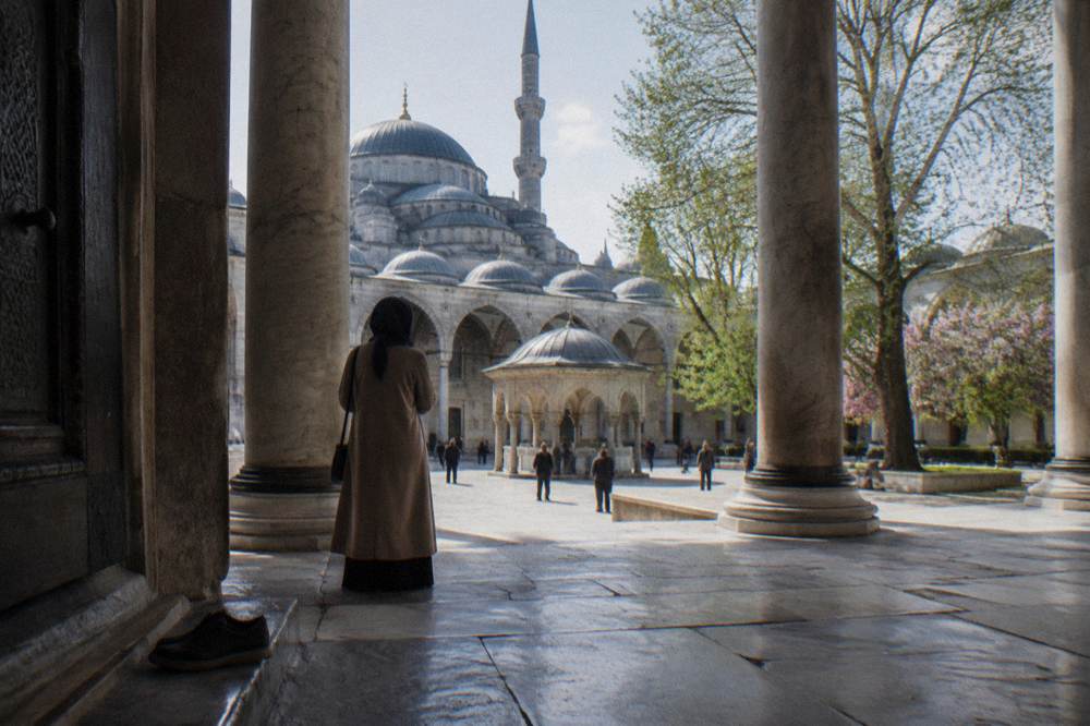 Visitor in a long coat and headscarf standing in the colonnaded courtyard of a large Ottoman mosque